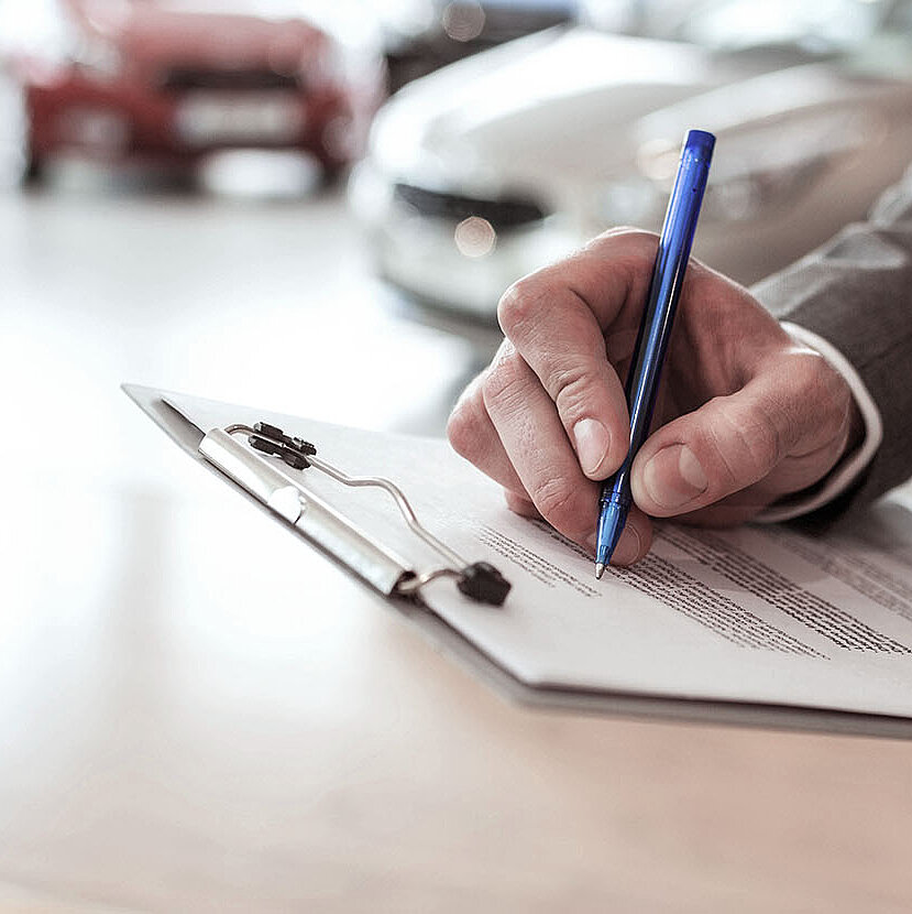 Eine Person im Anzug schreibt mit einem blauen Stift auf einem Klemmbrett, im Hintergrund sind unscharfe Autos zu sehen, was auf ein Autohaus oder ein Automobilumfeld schließen lässt.