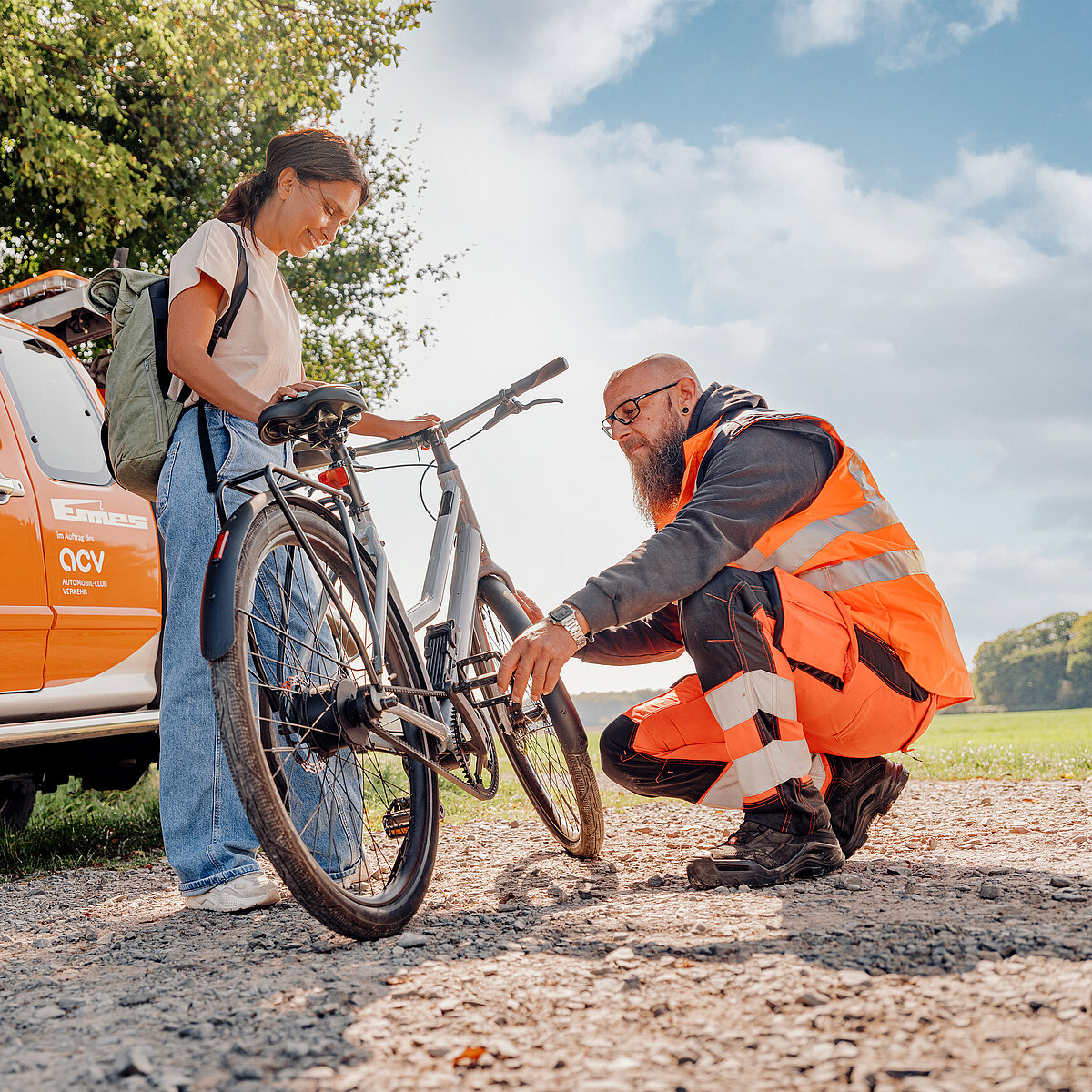 „Pannenhelfer in Warnweste repariert ein Fahrrad, während die Besitzerin daneben steht; im Hintergrund ein ACV-Servicefahrzeug.“
