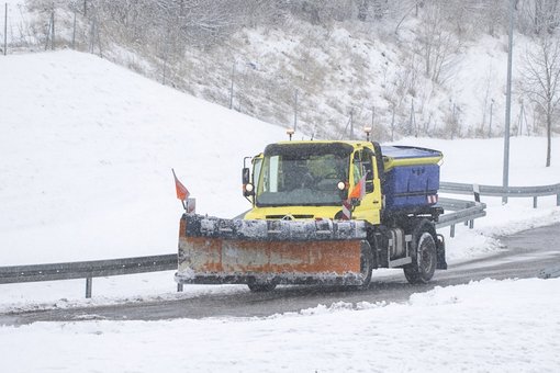 Ein Schneepflug fährt während starkem Schneefall über eine verschneite Straße. Das Fahrzeug ist gelb mit einem großen orangefarbenen Räumschild an der Front und einem blauen Streuaufsatz hinten. Der Schneepflug räumt Schnee von der Straße, während im Hintergrund schneebedeckte Hügel und Bäume sichtbar sind.