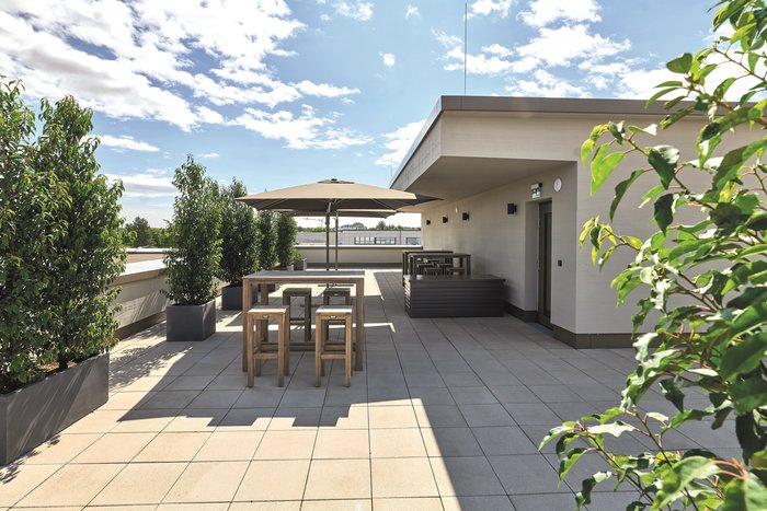 Dachterrasse mit modernen Sitzgelegenheiten, Holztisch und Stühlen, umgeben von Pflanzen und einer Pergola, unter einem sonnigen Himmel mit vereinzelten Wolken.