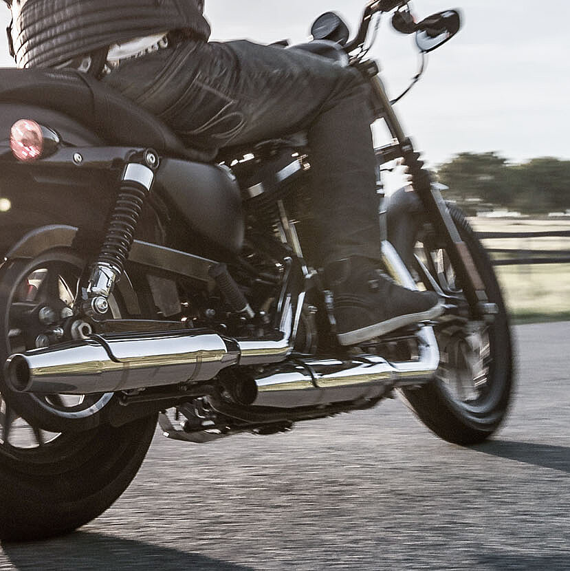Eine Person fährt auf einem schwarzen Motorrad auf einer asphaltierten Straße, wobei das Sonnenlicht die Szene und die grünen Felder im Hintergrund beleuchtet. Das Bild ist aus einem niedrigen hinteren Winkel aufgenommen, wodurch das Hinterrad und die Auspuffrohre des Motorrads betont werden.