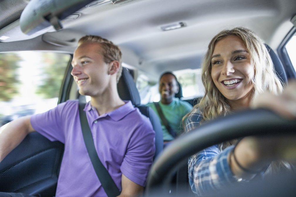 Eine junge Frau fährt ein Auto und lacht, während ein junger Mann auf dem Beifahrersitz ebenfalls lächelnd nach draußen schaut. Eine weitere Person sitzt auf der Rückbank. Das Bild symbolisiert eine fröhliche Fahrgemeinschaft und gemeinsames Reisen.