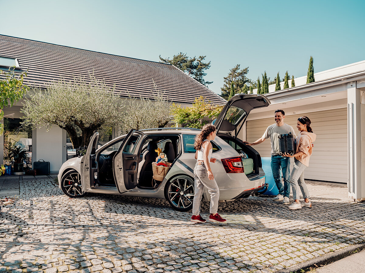 „Familie steht vor einem Haus und packt gemeinsam ihr Auto für die Reise, Kofferraum und Türen sind geöffnet.“