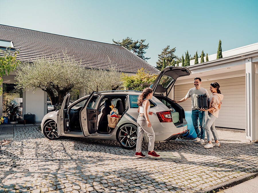 „Familie steht vor einem Haus und packt gemeinsam ihr Auto für die Reise, Kofferraum und Türen sind geöffnet.“