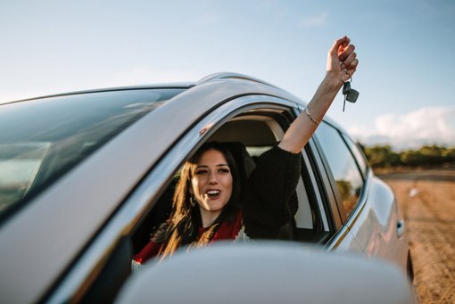 Junge Frau freut sich über ihr erstes eigenes Auto und hält stolz den Autoschlüssel aus dem Fenster