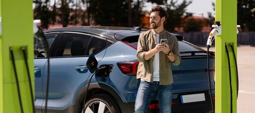 Ein Mann lehnt sich an ein blaues Elektroauto, während er es an einer öffentlichen Ladestation auflädt, hält sein Handy in der Hand und lächelt. Das Ladekabel ist an das Auto angeschlossen, und die Szene spielt draußen auf einem Parkplatz.