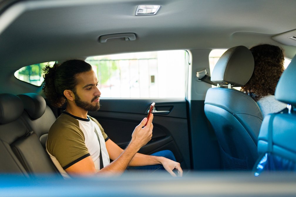 Ein Mann mit langen Haaren und Bart sitzt angeschnallt auf der Rückbank eines Autos und blickt auf sein Smartphone. Vorne sind die Fahrerin und eine weitere Person auf dem Beifahrersitz zu sehen. Das Bild symbolisiert eine Fahrgemeinschaft und moderne Mobilität.