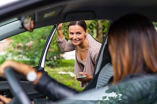 Mitfahren 2.0 Eine Frau mit einem Handy in der Hand steigt lächelnd in ein Auto ein, während die Fahrerin im Vordergrund auf sie wartet. Das Bild symbolisiert eine Fahrgemeinschaft und gemeinsames Reisen.