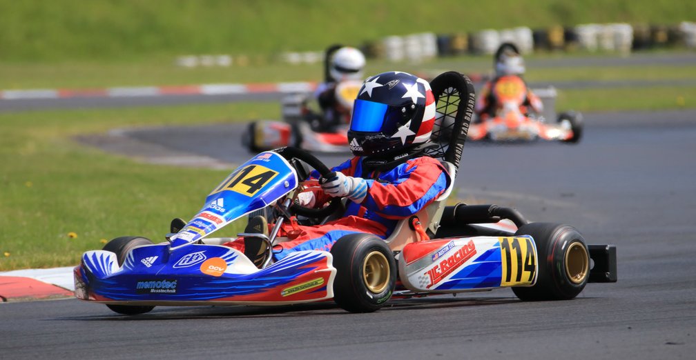 ACV German Karting Series GKS: Ein Fahrer in einem Rennkart, gekleidet in Schutzkleidung und Helm, fährt dynamisch durch eine Kurve auf einer Rennstrecke, begleitet von anderen Fahrern im Hintergrund