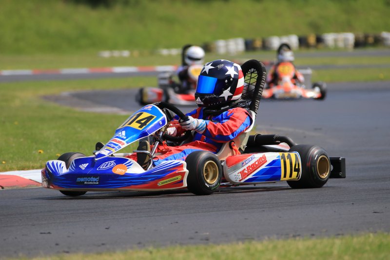 ACV German Karting Series GKS: Ein Fahrer in einem Rennkart, gekleidet in Schutzkleidung und Helm, fährt dynamisch durch eine Kurve auf einer Rennstrecke, begleitet von anderen Fahrern im Hintergrund