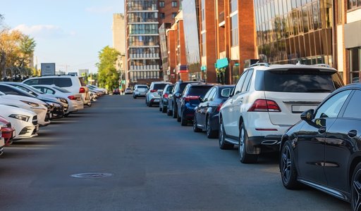 Eine belebte städtische Straße, gesäumt von parkenden Autos auf beiden Seiten. Im Hintergrund sind moderne Gebäude und eine teilweise bewölkte, sonnige Umgebung zu sehen.