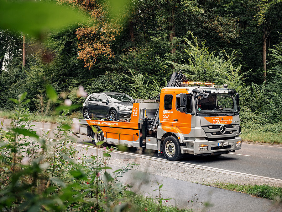 „Orangefarbener ACV Abschleppwagen fährt auf einer Landstraße, ein graues Auto ist auf der Ladefläche transportiert.“