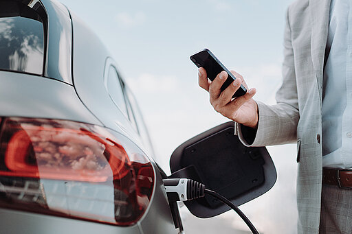 Mann steht mit seinem Handy in der Hand vor einem Elektroauto das lädt