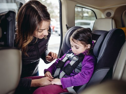 Das Bild zeigt eine Frau, die sich liebevoll um ein kleines Kind kümmert, das in einem Kindersitz im Auto sitzt. Die Frau befestigt oder überprüft den Sicherheitsgurt des Kindes, während sie sich leicht zu ihm hinüberbeugt. Das Kind trägt eine violette Jacke und sitzt sicher in einem gut gepolsterten Kindersitz. Im Hintergrund sind Teile des Autoinnenraums und das Fenster zu sehen, durch das Tageslicht hereinfällt. Die Szene vermittelt eine fürsorgliche und sichere Atmosphäre.