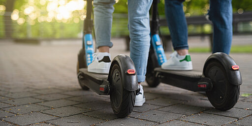 blackday - stock.adobe.com Zwei Personen in Jeans und Turnschuhen fahren mit einem Elektroroller auf einem gepflasterten Weg im Freien, mit grünen Bäumen und Sonnenlicht im Hintergrund. Das Bild ist von hinten aufgenommen und konzentriert sich auf ihre Beine und Roller.