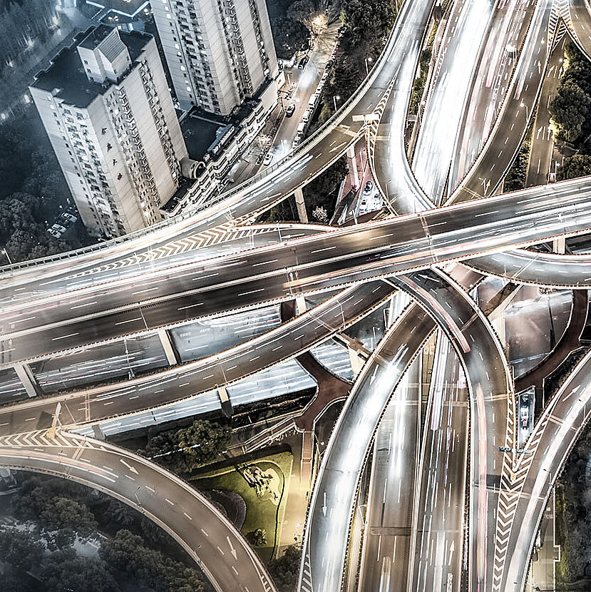 Luftaufnahme eines komplexen mehrstufigen Autobahnkreuzes mit Lichtspuren von fahrenden Fahrzeugen, umgeben von Gebäuden und Grünflächen.