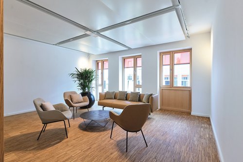 Heller Lounge-Bereich mit modernen Sesseln, Sofa, rundem Couchtisch und einer großen Zimmerpflanze, vor großen Fenstern, die natürliches Licht hereinlassen.