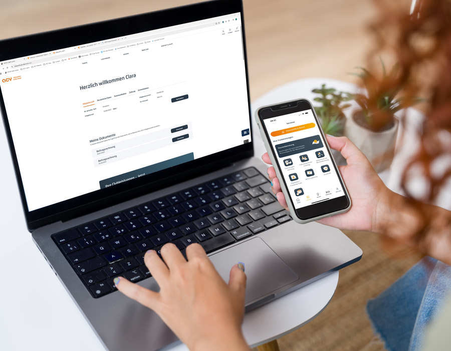 „Frau arbeitet an einem Laptop und hält ein Smartphone mit geöffneter ACV-App in der Hand.“