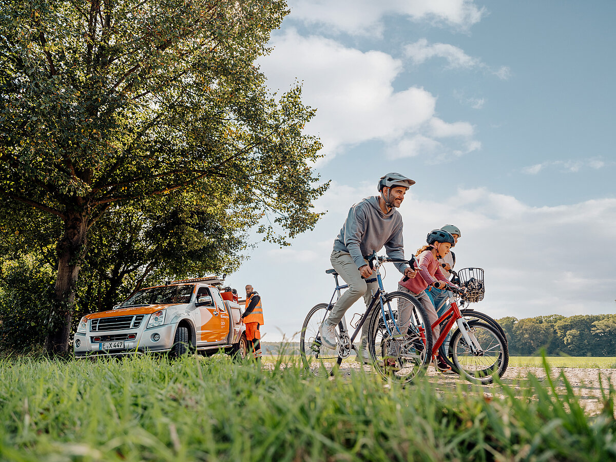 „Vater und Kind fahren mit Fahrrädern über einen Feldweg, im Hintergrund steht ein ACV-Servicefahrzeug mit Pannenhelfer.“