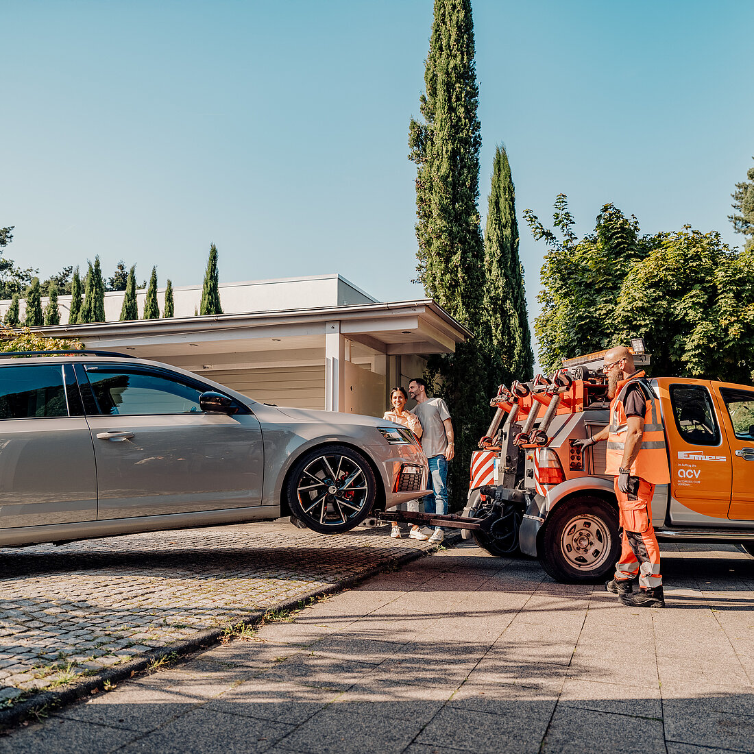 „Pannenhelfer lädt ein silbernes Auto mit angehobenen Vorderrädern auf einen orangefarbenen Abschleppwagen, während ein Paar zusieht.“