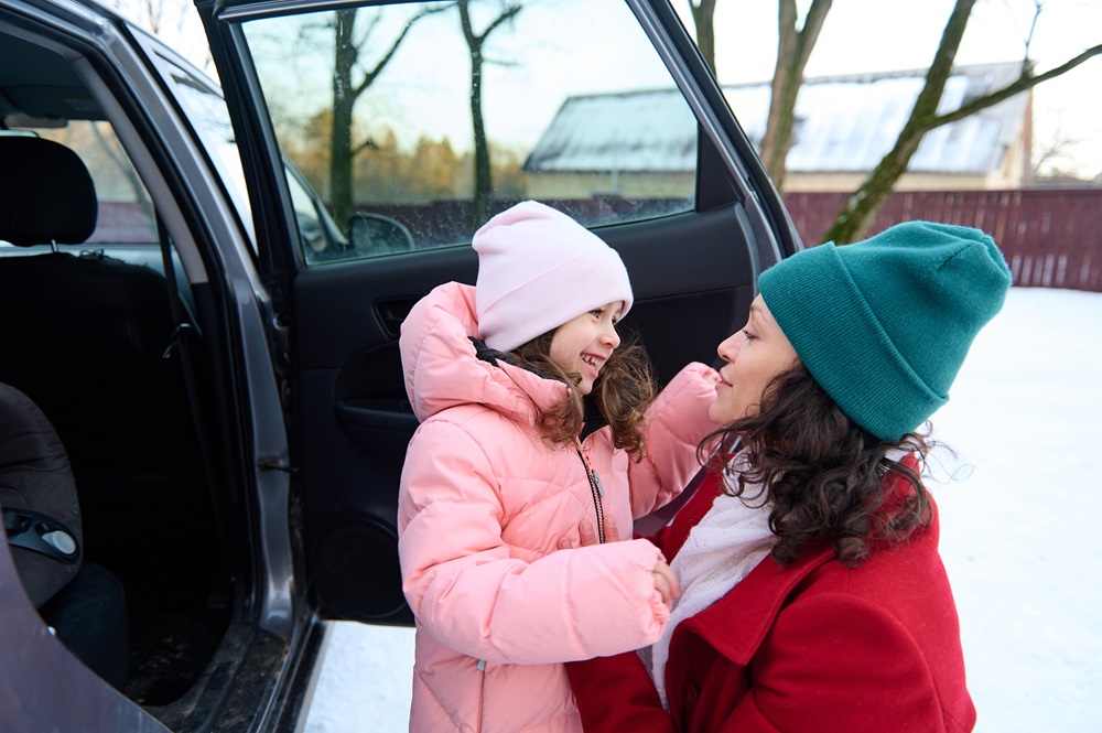 Das Bild zeigt eine Frau und ein Kind, die draußen in einer winterlichen Umgebung stehen. Die Frau trägt eine rote Jacke und eine grüne Mütze, während das Kind eine rosa Jacke und eine weiße Mütze trägt. Sie stehen in der Nähe eines Autos mit geöffneter Tür, und die Frau scheint fürsorglich das Gesicht des Kindes zu berühren oder etwas anzupassen. Im Hintergrund sind schneebedeckte Bäume und ein Zaun zu sehen, was auf eine kalte, winterliche Atmosphäre hinweist.