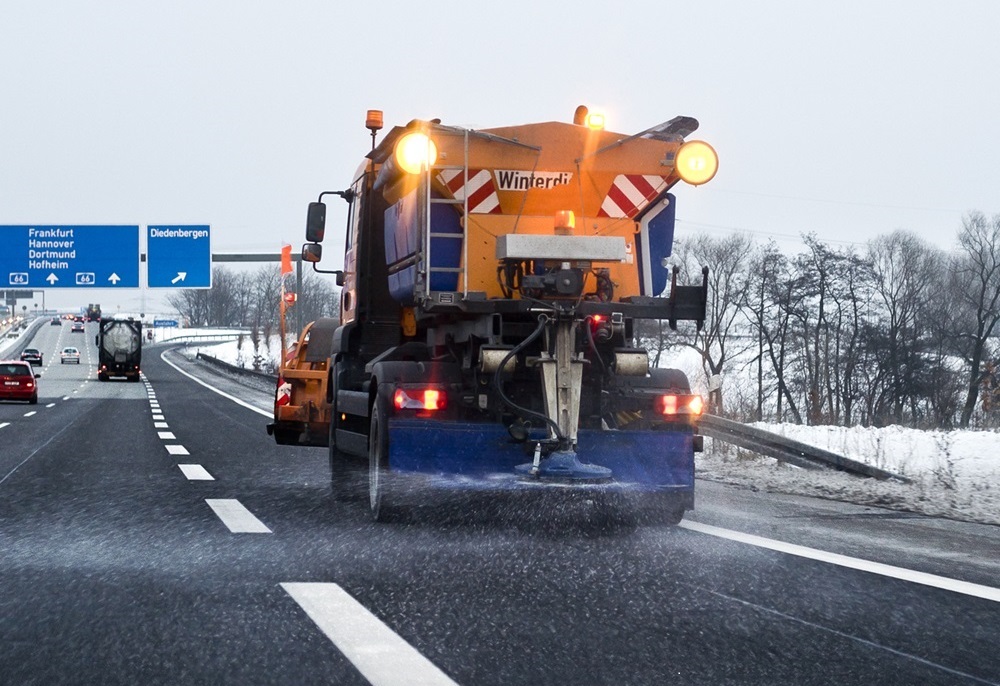 Ein Schneepflug fährt auf einer Autobahn und streut Streusalz auf die Fahrbahn. Das orangefarbene Fahrzeug ist von hinten zu sehen, mit einem blauen Streuer im Einsatz. Die Fahrbahn ist nass und teilweise von Schnee oder Eis bedeckt, während der Himmel grau und winterlich erscheint. Im Hintergrund sind weitere Fahrzeuge auf der Autobahn unterwegs, und Bäume stehen am Straßenrand.