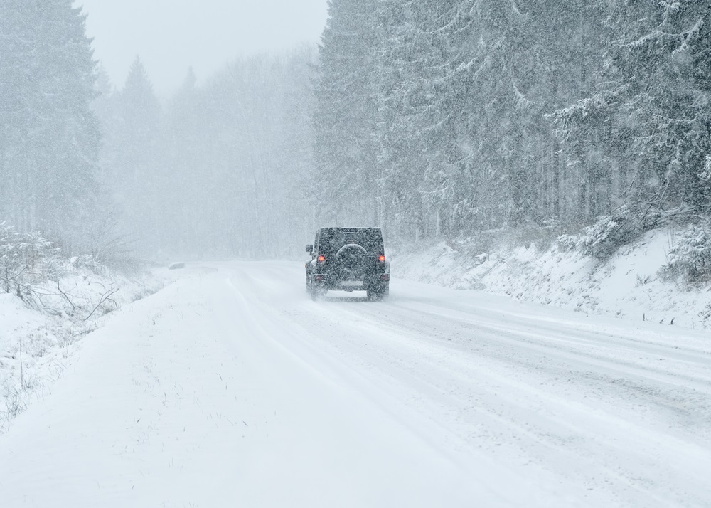 Auto fährt auf einer verschneiten Landstraße durch dichten Schneefall, umgeben von schneebedeckten Bäumen. Winterliche Straßenverhältnisse und Autofahren bei Schnee.