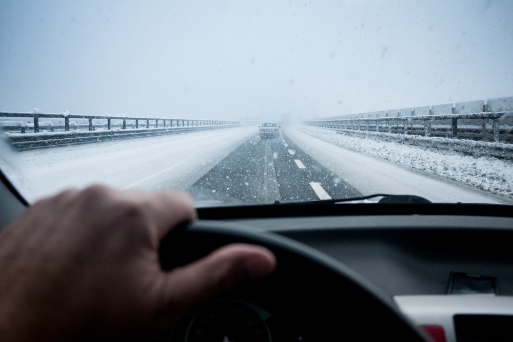 Eine winterliche Straße, bei der der Fahrer durch die Windschutzscheibe eine schneebedeckte Straße sieht. Die Fahrbahn ist rutschig, und der umliegende Schnee weist auf schwierige Fahrbedingungen hin