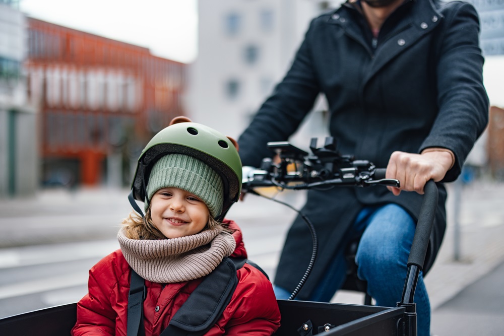 Das Bild zeigt ein fröhliches Kind, das in einer Transportbox eines Lastenfahrrads sitzt. Das Kind trägt eine rote Jacke, einen grünen Helm und einen dicken Schal, passend zur kühlen Jahreszeit. Hinter dem Kind ist ein Erwachsener in dunkler Kleidung zu sehen, der das Fahrrad lenkt. Der Hintergrund zeigt eine städtische Umgebung mit Gebäuden und Straßen, was auf eine urbane Fahrradtour hinweist. Die Szene vermittelt eine warme und sichere Atmosphäre.