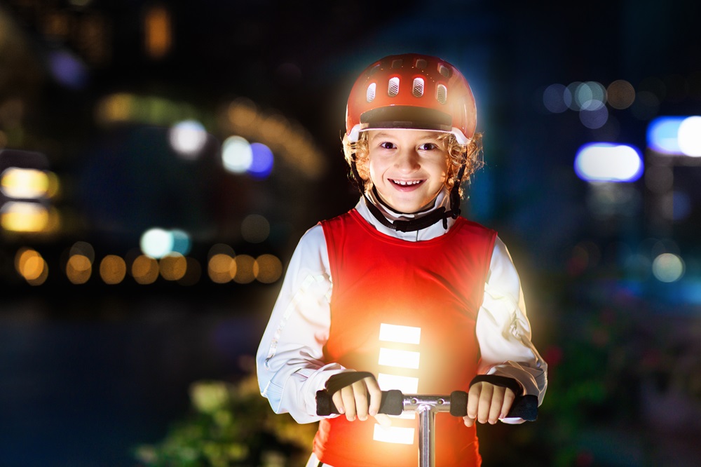 Das Bild zeigt ein fröhliches Kind, das im Dunklen auf einem Roller unterwegs ist. Das Kind trägt eine rote Sicherheitsweste und einen passenden Helm, während ein helles Licht am Fahrzeug für gute Sichtbarkeit sorgt. Im Hintergrund ist eine nächtliche Umgebung mit unscharfen Lichtern zu erkennen, was auf eine städtische oder beleuchtete Umgebung hinweist. Die Szene vermittelt Sicherheit und Freude am nächtlichen Fahren.