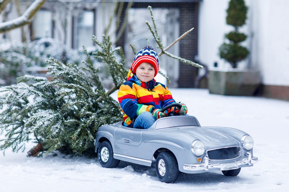 Ein lachender Junge in einem farbenfrohen Winteroutfit sitzt in einem kleinen Spielzeugauto auf verschneitem Boden, während hinter ihm ein Weihnachtsbaum am Auto befestigt ist. Die Szene spielt in einem winterlichen Vorgarten.