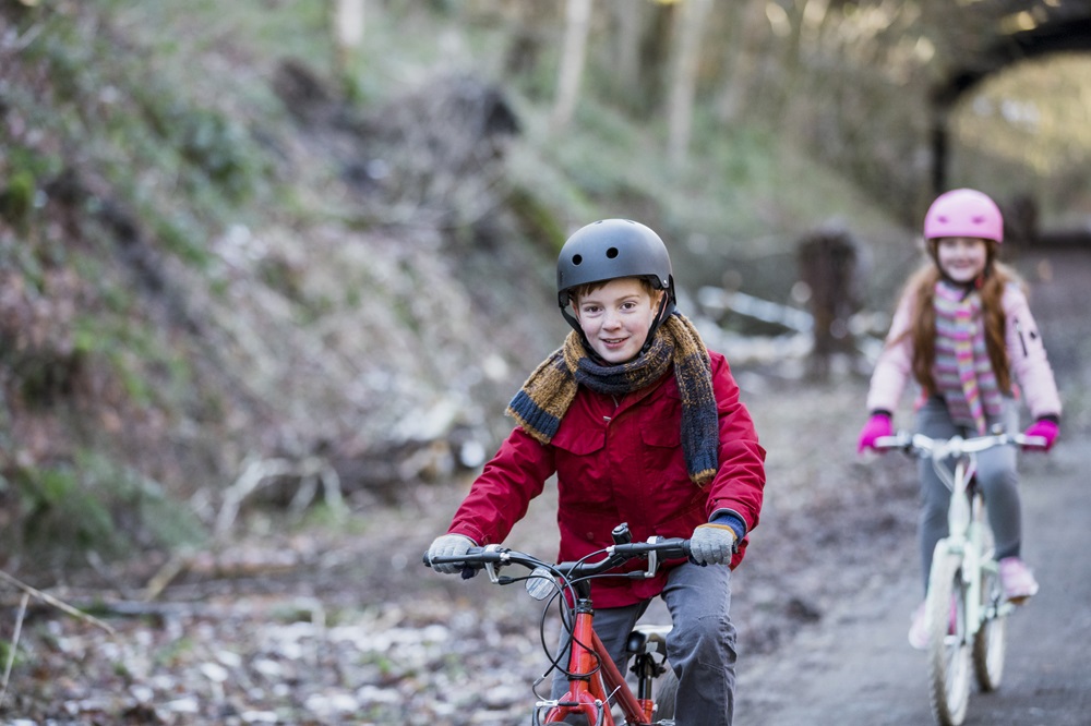 Das Bild zeigt zwei Kinder, die auf Fahrrädern auf einem Weg durch eine naturnahe Umgebung fahren. Das vordere Kind trägt eine rote Jacke, einen schwarzen Helm und fährt ein rotes Fahrrad. Das hintere Kind trägt eine rosa Jacke, eine passende Mütze und fährt ein weißes Fahrrad. Beide Kinder scheinen sich an einem kalten Tag draußen zu bewegen, und im Hintergrund sind Bäume und ein ruhiger, natürlicher Pfad zu sehen. Die Szene wirkt aktiv und fröhlich.