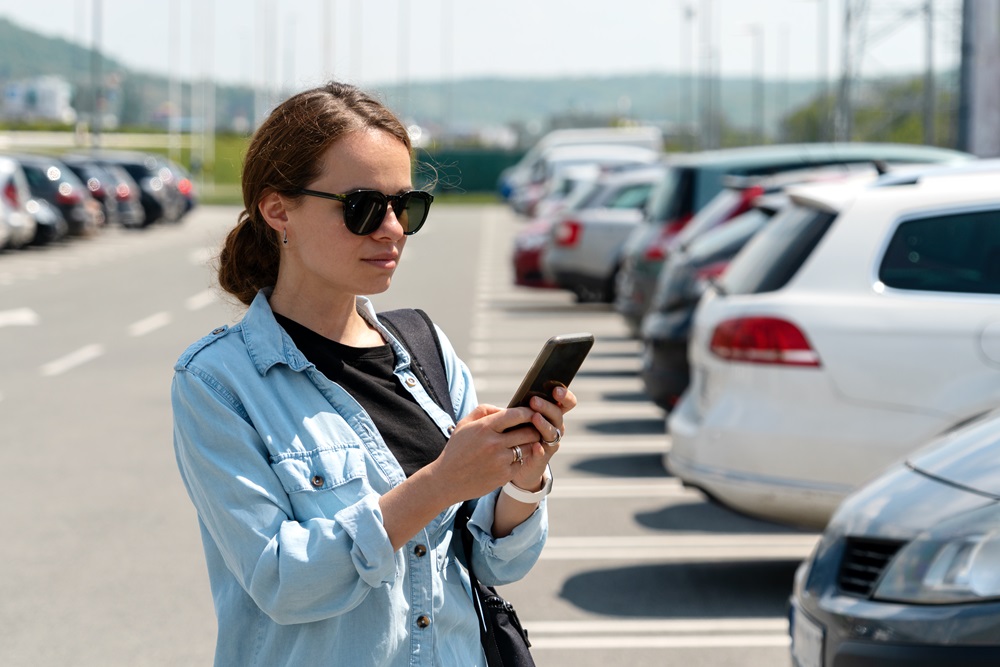 Eine Frau mit Sonnenbrille und lässiger Kleidung steht auf einem Parkplatz und schaut auf ihr Smartphone. Im Hintergrund sind mehrere parkende Autos und eine ländliche Umgebung mit Hügeln zu sehen.