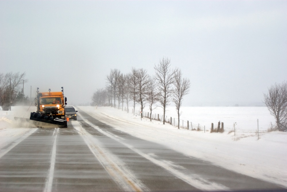 Ein Schneepflug fährt auf einer winterlichen, teils geräumten Landstraße. Der gelb-orangefarbene Schneepflug befindet sich auf der linken Seite der Fahrbahn und räumt Schnee. Die Straße führt durch eine schneebedeckte Landschaft mit kahlen Bäumen am Straßenrand und Feldern, die ebenfalls von Schnee bedeckt sind. Der Himmel wirkt trüb und winterlich.