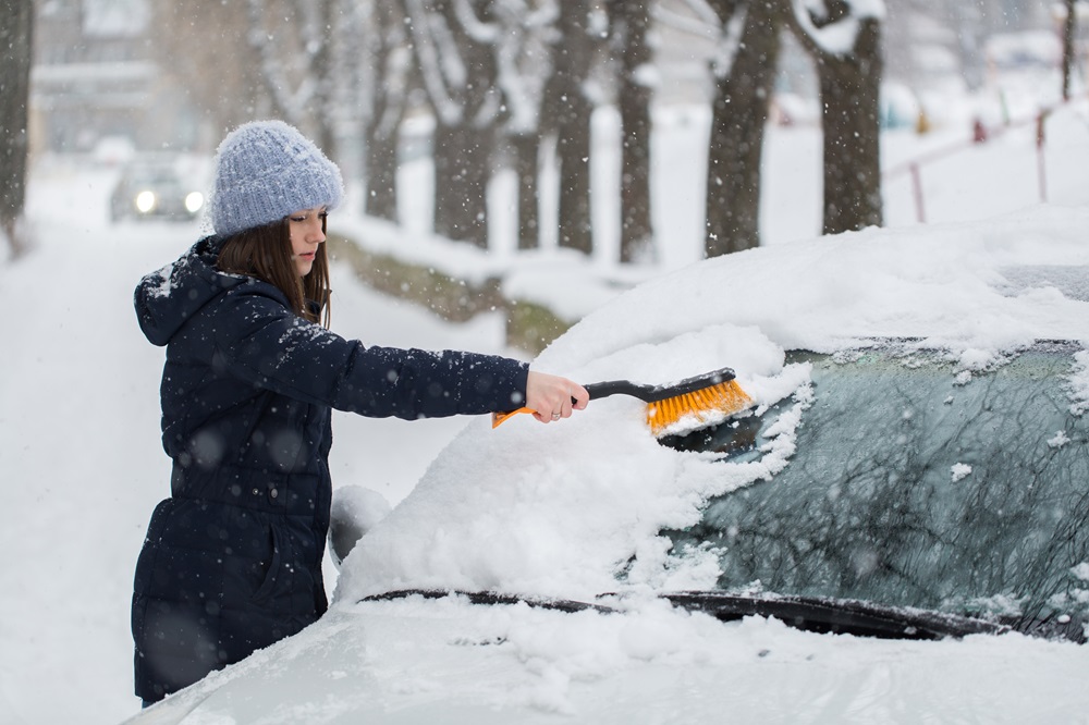 Frau in Winterkleidung entfernt Schnee von der Windschutzscheibe eines Autos mit einem Schneebesen auf einer verschneiten Straße