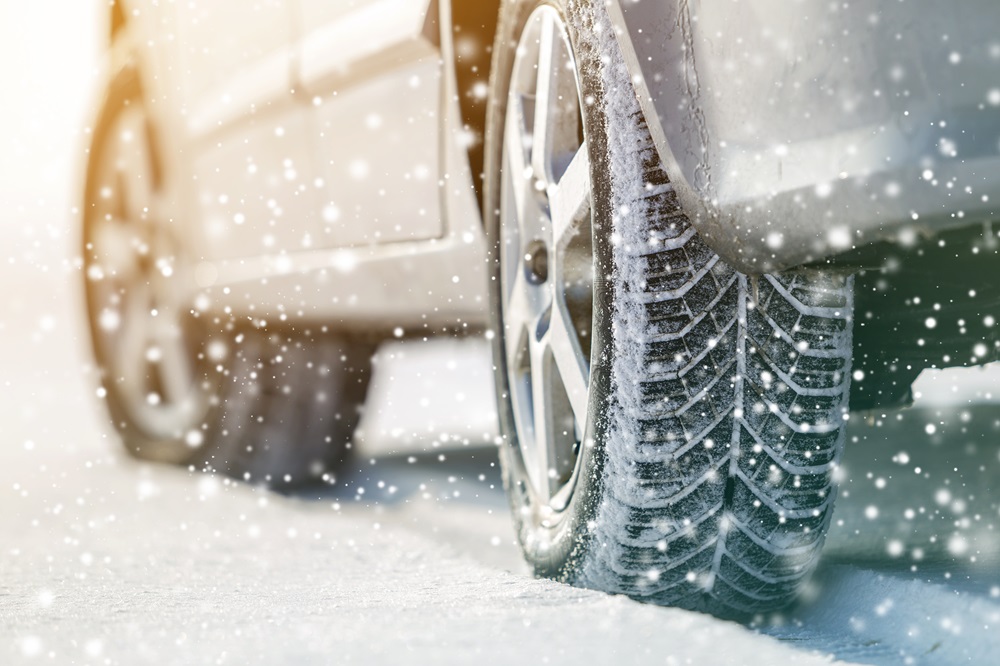 Detailaufnahme eines Autoreifens mit Winterprofil auf einer verschneiten Straße, umgeben von fallendem Schnee. Sicherheit beim Fahren im Winter und Winterreifen.