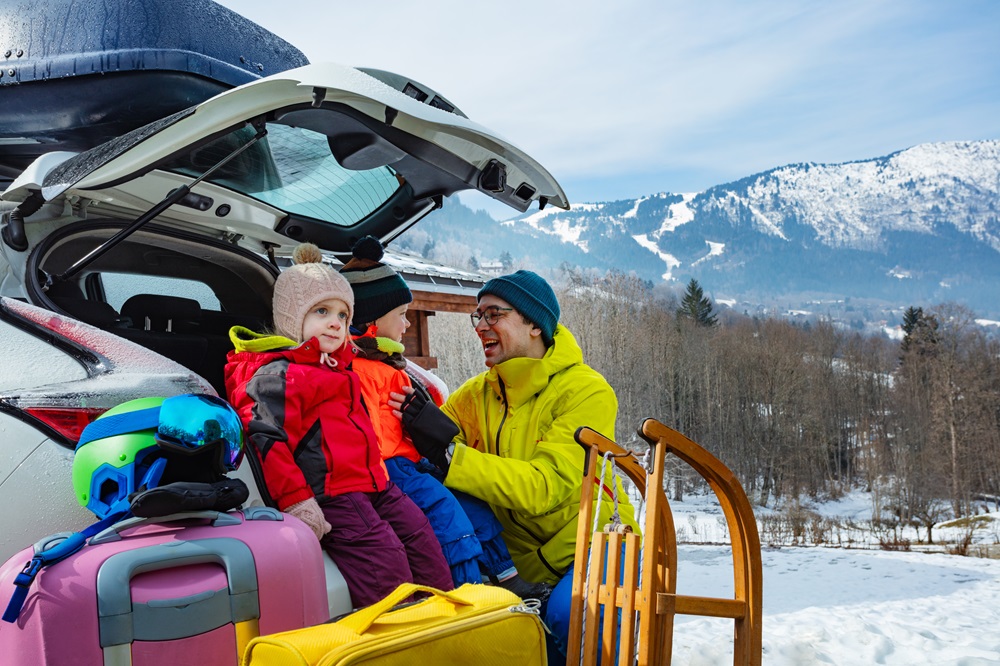 Eine Familie bereitet sich auf einen Winterurlaub vor. Im Vordergrund sind bunte Koffer und Taschen zu sehen, während ein Erwachsener einem Kind mit roter Jacke und Mütze hilft, sich warm anzuziehen. Im Hintergrund erstreckt sich eine schneebedeckte Berglandschaft unter einem strahlend blauen Himmel, was die winterliche Atmosphäre unterstreicht.